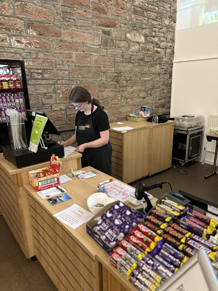 A volunteer with glasses standing centre-right of the picture wearing The Usual Place uniform. She is stood behind the welcome station using the register.