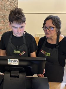 A volunteer with brown hair standing to the left of the photo wearing The Usual Place uniform. Standing next to him is a mentor with glasses who is also wearing The Usual Place uniform. They are looking at the welcome station's till screen, concentrated on putting the customer's order into the system.