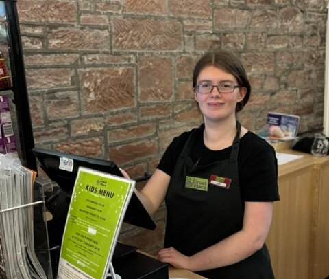 Young woman stands behind a till and smiles. She is wearing a black uniform with an apron, glasses and has her hair tied back