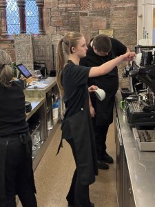 A volunteer with blonde hair standing centre of the photo and another volunteer with brown hair stood behind her. They are working at the Drinks and Cakes station, preparing orders for the customers. To the right of the photo is a mentor. They are all wearing The Usual Place uniforms.