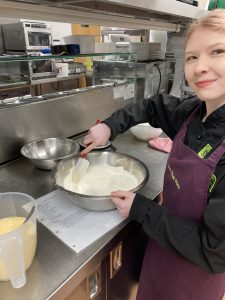 On the far right-hand side of the photo, a young woman/girl is looking at the camera and smiling. She is wearing a purple apron with 'The Usual Place' logo emblazoned on it. She is holding a white spatula in her right hand, which she is using to stir cake mix in a metal bowl, the rim of which she is holding in her left hand. Underneath the bowl is a recipe which she is following, and to the left of that is a jug containing a mix. Above the cake bowl is another empty metal bowl that is slightly smaller.