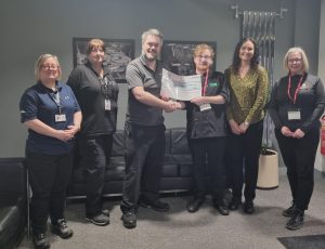 A volunteer stands with team members from Gates Dumfries and a mentor from The Usual Place. To the left of the photo are three of the team members, one of which the volunteer is holding a certificate of donation, and to the right of the photo is another team member and The Usual place mentor. The volunteer and mentor are wearing The Usual Place uniform and building passes around their necks. Everyone is looking into the camera and smiling.