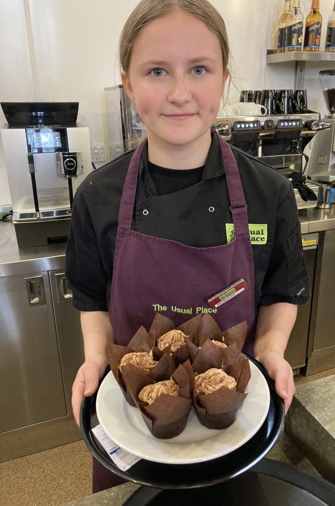 A girl is in the centre of the photo, wearing a purple apron and a name badge. The words 'The Usual Place' are emblazoned on the apron in green. The girl is smiling and looking straight at the camera, and she is holding a black platter, on top of which is a white plate, that has five chocolate muffins on it. Behind her is a workstation for making drinks.