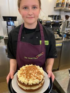 A girl is in the centre of the photo, wearing a purple apron and a name badge. The words 'The Usual Place' are emblazoned on the apron in green. The girl is smiling and looking straight at the camera, and she is holding a black platter, on top of which is a white plate, that has a sticky toffee cake with cream-coloured icing on it, decorated with caramel chunks and drizzle on it. Behind her is a workstation for making drinks. 