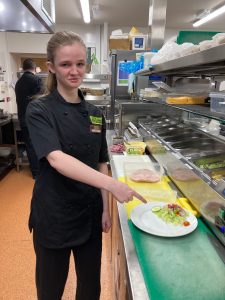 A girl is stood towards the left of the photo in a black kitchen uniform, which has 'The Usual Place' logo emblazoned on it on the viewer's right, and she is wearing a name badge underneath it. She is smiling, looking at the camera, and pointing with her right hand, at a plate with a salad on it, which is sat on a green chopping board which is on a metal counter top. directly above (to the viewer's perspective), there is a yellow chopping board with cooked meat in a tub on it. To the right of the chopping boards there is a row of embedded metal containers which have various different toppings inside them.