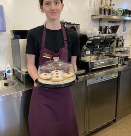 A volunteer with brown hair standing in the centre of the photo wearing The Usual Place uniform. She is smiling and holding a container of biscuits next to the barista machine.