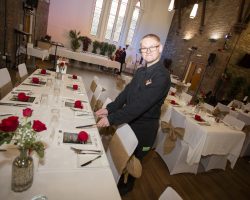 Ryan prepares tables before the guests arrive young man smiles at the camera whilst setting a long table in a highly decorated style in an event space