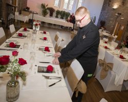 Ryan concentrates on preparing tables before our guests arrive Youn gman wearing red glasses stands behind a highly decorated table with an expression of concentration whilst he ensure the knives are carefully lined up