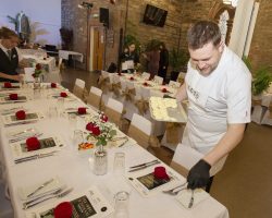 Chef Fraser Cameron ensures every guest has a perfectly formed butter when they arrive!. A chef dressed in whites with 1610 logo and gloves is standing behind a highly decorated table and carefully placing small formed butter pats on the side plates