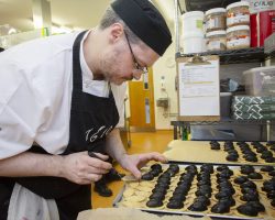 Chef Garry prepares seafood accompaniment to the Salmon Wellington Starter in The Usual Place kitchen Chef in white jacket, black cap and apron prepares many delicate pastries with black cream toppings in a shiny kitchen.