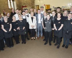 Team photo just before the doors open to guests. Outside, Piper James Gray entertains the waiting guests. A team of people face the camera and smile. In the centre are the two comperes and they are surrounded by chefs and other team members dressed in black with aprons and black gloves.