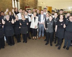 A final cheer before we open the doors and the whole team moves into action to welcome guests and plate up. A team of smiling people face the camera and raise their hands in joy. In the centre are the two comperes and they are surrounded by chefs and other team members dressed in black with aprons and black gloves