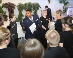 Hospitality Manager, Chitra, gives final instructions before we open the doors Man dressed in a suit, with black service gloves and a sheet of notes gives motivational instructions to the team of people standing around him and listening intently.