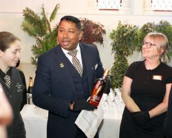Chitra introduces some of the drinks including a fine whisky from Annandale Distillery which guests will enjoy for the Sante du Chef at the end of the event. Man in suit and tie with service gloves holds a bottle of whisky. Other members of the team listed to his instructions and everyone is smiling in anticipation of the event starting.