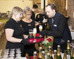 Gail, Josephine and Richard prepare the Corney & Barrow Sparkling to welcome guests as they arrive Three people stand around a tray that one of them is holding and another is adding glasses of sparkling wine. In front of them are lots of bottles and glasses lined up neatly and in the back ground other members of the team are welcoming guests.