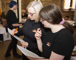 Sharon and Leah make sure they are ready to guide each guest to their correct table. Two women stand holding paper and pens, they are discussing the details.