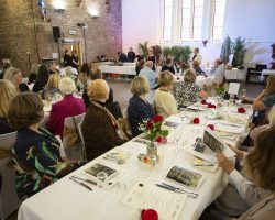 Fiona Armstrong welcomes everyone and introduces her co-host Lewis Shaw A view across the room from behind a long table with seated guests. At the far end a lady dressed in a white jacket is standing and speaking into a microphone. Behind her is a drinks service area.
