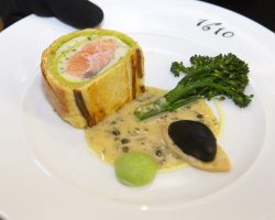 The starter: Salmon Wellington, Seafood Cream, Broccoli and Champagne Butter Sauce A beautifully presented plate of food. The plate is white and has the logo 1610 printed on it. Also on the plate is salmon wrapped in watercress mouse and pastry, a creamy sauce, a spear of broccoli and two side items which are green and black. The plate is being held by a server wearing black gloves.