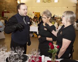 Making sure everyone is ready to serve the wine pairings with each course. The Bar Manager and two servers are discussing wine pairings. In front of them are wine glasses on trays as well as red rose display. IN the background are the tables full of guests.