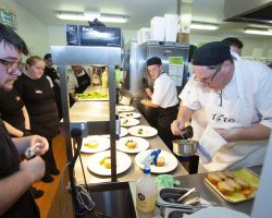 A view from the side of the hot pass in the kitchen. On the right are the chefs who are plating up, in the middle are the plates and on the left are the servers standing waiting for the go-ahead to pick up the plates to take to the guests.