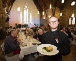 Ryan with our salmon wellington starter A young waiter stands holding a plate of beautifully presented food. In the background are tables with many guests all enjoying eating and socialising.