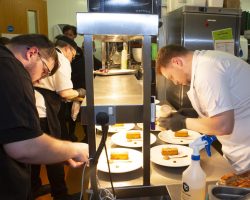 Chefs on both sides of the hot pass are plating and checking each plate of food as the waiting team look on ready for instruction to begin serving to guests.