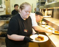 Young waitress takes a plate of beautifully prepared food and is about to leave the kitchen, behind her a chef is ensuring each plate is well presented and ready for serving.
