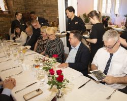 Guests sit at a table as waiters and waitresses begind to bring the main course dishes behind them. The guests are chatting and reading the menu
