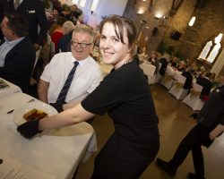 A young waitress smiles at the camera as she places a plate of food in front of CEO Craig McEwen at a table. In the background lots of other guests at tables are enjoying their main course.