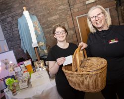Leah and Sharon prepare to draw the raffle. Two ladies stand in the foreground with a wicker basket. IN the background are raffle prizes and gifts including a pale blue silk jacket made from kimono silk