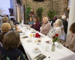 Singer Claire Hastings entertains during coffee A young woman stands and sings with a ukelele in the background. In the foreground guests sit at tables listening and enjoying the music.