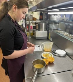 A volunteer with brown hair stood to the left of the photo wearing The Usual Place uniform. She is standing in front of the kitchen counter and making food.