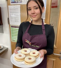 A volunteer with brown hair standing in the centre of the photo wearing The Usual Place uniform. She is holding a plate of biscuits and smiling into the camera.