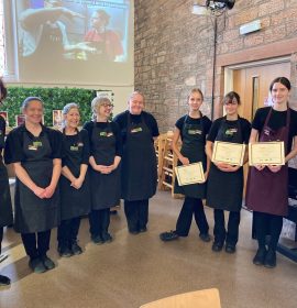 3 volunteers lined up standing to the right of the photo, holding their certificates next to a mentor and smiling. On the right are 5 other mentors lined up and smiling with them. They are all wearing The Usual Place uniforms.