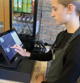 A volunteer with blonde hair standing to the right of the photo. She is looking at the screen while taking a customer's order at the till.
