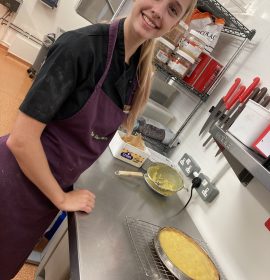 A volunteer with blonde hair standing to the left of the photo wearing The Usual Place uniform. She is smiling and standing in front of the kitchen counter which displays a pie she has baked.
