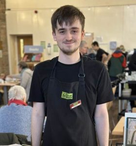 A volunteer standing centre of the photo wearing The Usual Place uniform. He is smiling and standing behind the Welcome board, ready to greet customers and show them to their tables.