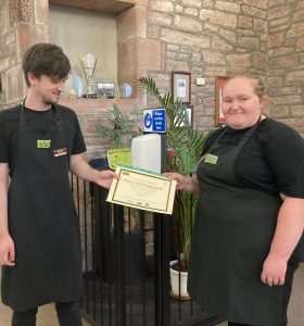 A volunteer standing to the left of the photo and a mentor standing to the right of the photo wearing The Usual Place uniform. They are holding the volunteer's certificate and smiling.