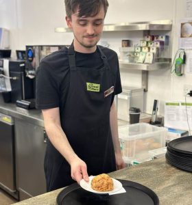 A volunteer standing centre-left of the photo wearing The Usual Place uniform. He is standing at the Cakes And Drinks station counter, about to place a plate with a scone onto a tray for someone to take to a customer.