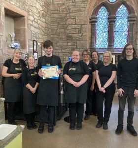 8 people standing centre of the photo wearing The Usual Place uniform. One is a volunteer holding his certificate for completing his training, while the others are mentors and staff standing next to or behind him. They are all looking at the camera and smiling.