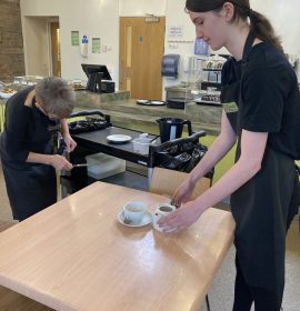 A volunteer with brown hair standing to the right of the photo wearing The Usual Place uniform. She is holding a mug and a plate, clearing the table. Behind her is a black trolley with used plates on it and a mentor helping her clean the table for the next customer.