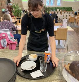 A volunteer with glasses bending to pick up a tray off of the counter. The tray has an assortment of coffee and drinks placed on it, ready to be taken to the customer.