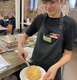 A volunteer with glasses standing centre-right of the photo wearing The Usual Place uniform. She is smiling and preparing a scone to put onto the tray and send out to the customer.