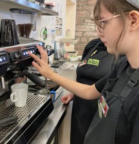A volunteer with glasses standing to the right of the photo. She is using the barista machine and making a coffee for a customer.