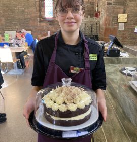 A volunteer with glasses standing in the centre of the photo wearing The Usual Place uniform. She is smiling and holding a container with a cake inside of it.