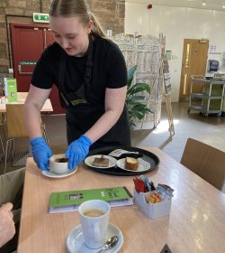 A volunteer with blonde hair standing centre-left of the photo wearing The Usual Place uniform and blue gloves. She is serving a customer their coffee and cake with a smile on her face.