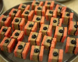 Canapes: Duck Rillette Sandwich and Black Garlic Emulsion
Whisky Smoked Salmon and Potato Salad Pumpkin & Mascarpone Gougère A close up of a tray of canapes. Each is a sandwich making up a small square with pink slices outside and a pate inside with a dot of black garlic emulsion in the middle. It looks very neat and every piece is exactly the same.