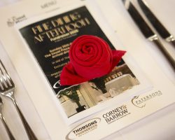 The Menu and the sponsors: Thomsons Food Services, Corney & Barrow and Patersons Fresh Fruit & Veg. Photograph of a place setting at a guest table, There is a white linen cloth, on top are layered a menu with the sponsors' logos presented. A printed programme sits on top and a red napkin, rolled into the shape of a rose is placed on top.