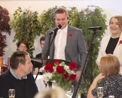 Lewis took our guests through every emotion from tears to laughter and back again with his insights and amazing compering skills. A young man dressed in a grey suit stands behind a mic and speaks. He has a hint of mischievous humour on this face. In the foreground guests are sitting at tables listening to him amongst table displays of red roses.