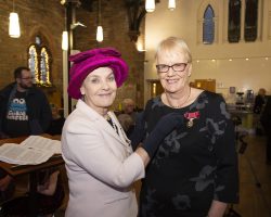 Smartly dressed lady in pink hat stands next to a tall middle-aged lady with short blonde hair and glasses. The former is wearing black gloves and is pinning a British empire Medal with red ribbon to the blonde lady's black dress